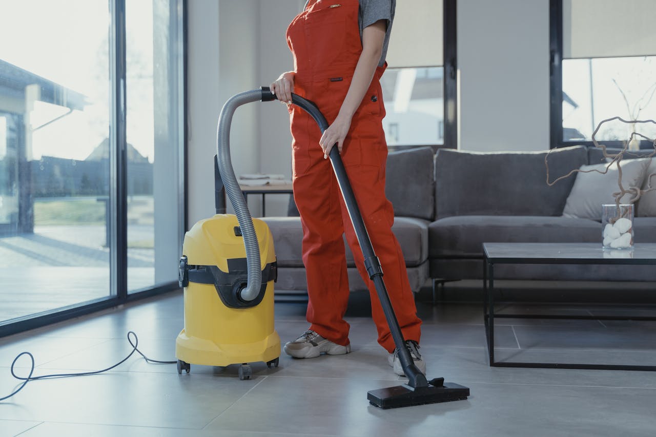 services-01 A professional cleaner in red coveralls vacuuming a modern living room with a yellow vacuum cleaner.